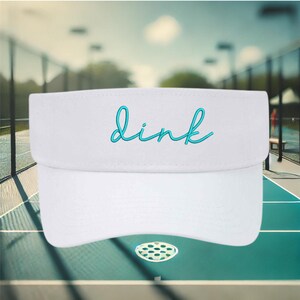 May include: White visor with a turquoise blue embroidered word "dink" on the front. The visor is on a green and white tennis court.