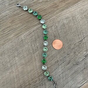 May include: A silver bracelet with a graduated ombre of green crystal beads. The bracelet is shown next to a US penny for size reference.