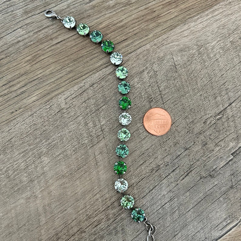 May include: A silver bracelet with a graduated ombre of green crystal beads. The bracelet is shown next to a US penny for size reference.