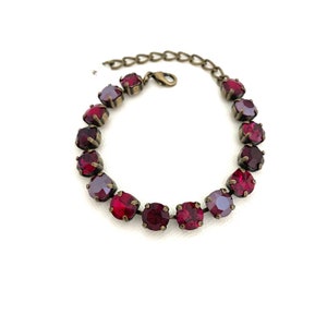 May include: A vintage-style bracelet featuring a series of round, deep red and purple-toned crystals. The crystals are set in a bronze-colored metal chain. The bracelet is displayed on a white background.