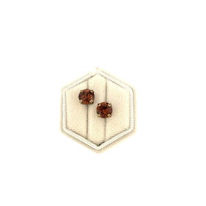 May include: A pair of stud earrings with round, reddish-brown gemstones set in gold-colored metal. The earrings are displayed on a cream-colored, hexagonal jewelry box with a soft, velvety interior.
