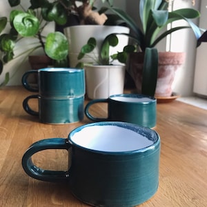 Może przedstawiać: Cztery tealowe ceramiczne kubki z białym wnętrzem. Kubki mają zaokrąglony kształt i uchwyt. Kubki są ułożone na drewnianej powierzchni.