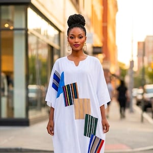 May include: A white floor-length kaftan dress featuring a V-neckline and short sleeves. The garment is decorated with several rectangular patches in various colours and patterns, including blue, orange, green, and multi-coloured vertical stripes.
