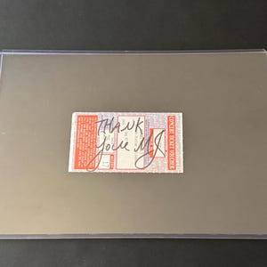 Puede incluir: Un vale de entrada para un concierto con detalles rojos y blancos, con texto manuscrito "Thank you, Jole MJ". El boleto está encerrado en un soporte de plástico transparente, sobre un fondo gris neutro.