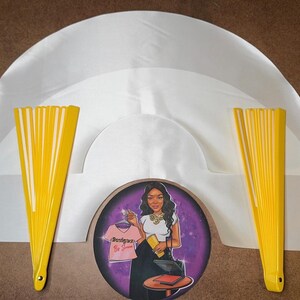 May include: Three white satin fabric fan shapes with yellow fabric fans on each side. The fans are closed and the fabric is smooth. The center fan shape has a round sticker with a woman in a purple background. The sticker says "Boss Lady" in pink letters.