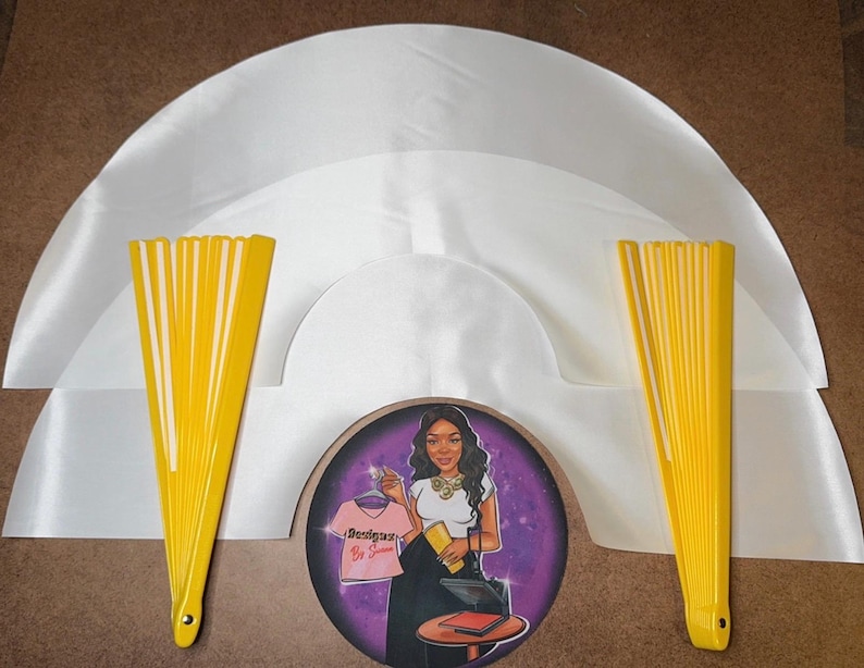 May include: Three white satin fabric fan shapes with yellow fabric fans on each side. The fans are closed and the fabric is smooth. The center fan shape has a round sticker with a woman in a purple background. The sticker says "Boss Lady" in pink letters.