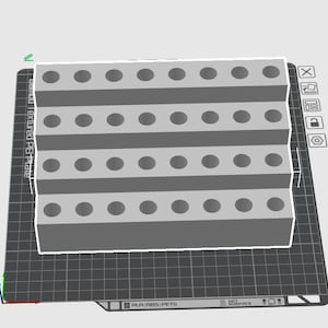 May include: A gray, tiered organizer with multiple circular holes, likely for holding tools or small items. The organizer sits on a textured, dark gray surface with a grid pattern. The design appears to be 3D-printed.