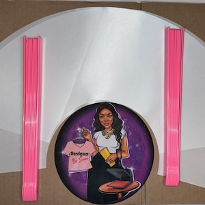 May include: A set of white, semi-circular, fabric garment protectors with pink plastic hangers. The protectors are arranged in a fan shape. A sticker with a cartoon image of a woman holding a t-shirt with the text "Designs by Simone" is in the center.