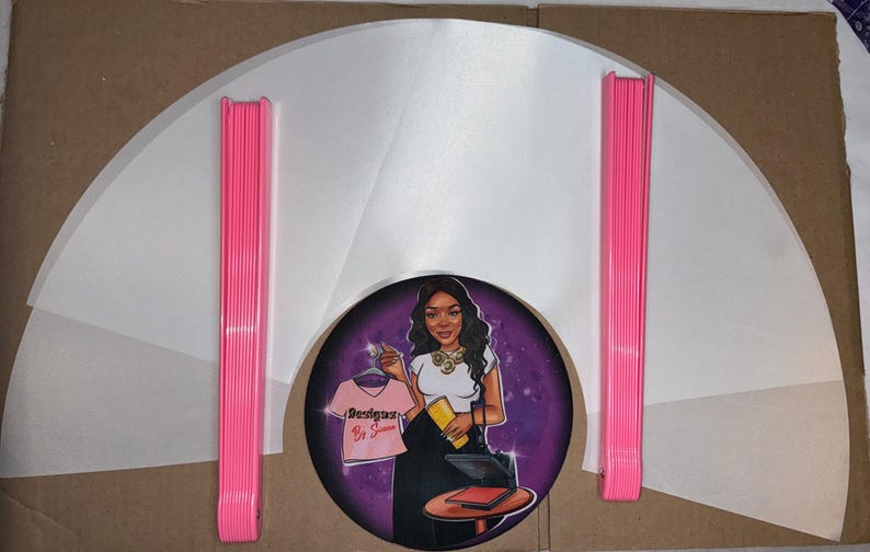 May include: A set of white, semi-circular, fabric garment protectors with pink plastic hangers. The protectors are arranged in a fan shape. A sticker with a cartoon image of a woman holding a t-shirt with the text "Designs by Simone" is in the center.