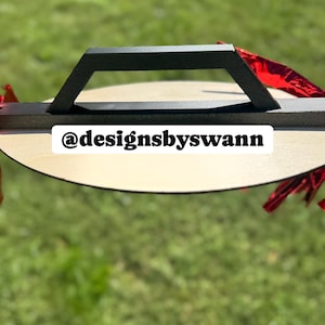 May include: A wooden serving tray with a black handle. The tray is round with a light wood finish. The text "@designsbyswann" is printed on the tray. Red decorative accents are visible.