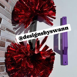 May include: Two red metallic pom-poms with black handles are displayed against a white wall. A purple wall hook is also visible. The text "@designsbyswann" is printed on a white banner.