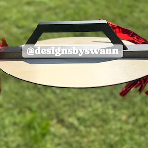 May include: A wooden serving tray with a black handle and the text "@designsbyswann". The tray has a light wood finish and is decorated with red accents. The background is blurred green grass.