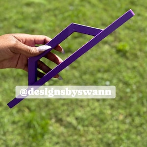 May include: A purple, geometric-shaped object with a rectangular frame and a diagonal arm. The object is held against a backdrop of green grass. The text "@designsbyswann" is visible.