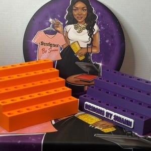 May include: Two tiered display stands for pens or markers, one in bright orange and one in purple. Each stand features four levels with multiple circular cutouts for storage. The purple stand has a white rectangular label reading Designs By Swann.