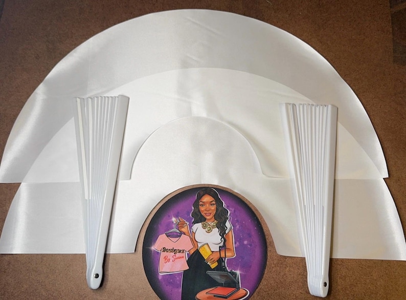 May include: Three white satin fabric fan shapes, two white paper folding fans, and a round sticker with a woman wearing a white shirt with the text "Designers By Jasmine" on it.