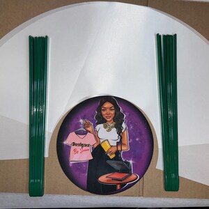 May include: A set of white paper fan shapes with green wire stems. The fan shapes are arranged in a semi-circle around a purple circle with a cartoon image of a woman holding a pink shirt with the text "Designer Be So Sweet" on it.