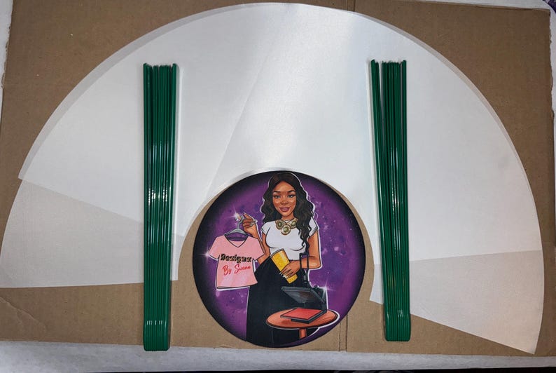 May include: A set of white paper fan shapes with green wire stems. The fan shapes are arranged in a semi-circle around a purple circle with a cartoon image of a woman holding a pink shirt with the text "Designer Be So Sweet" on it.