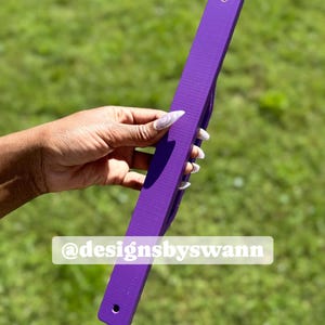 May include: A long, rectangular purple object with a textured surface and two holes, held against a green background. The text "@designsbyswann" is visible on the object. The object appears to be a ruler or a similar measuring tool.