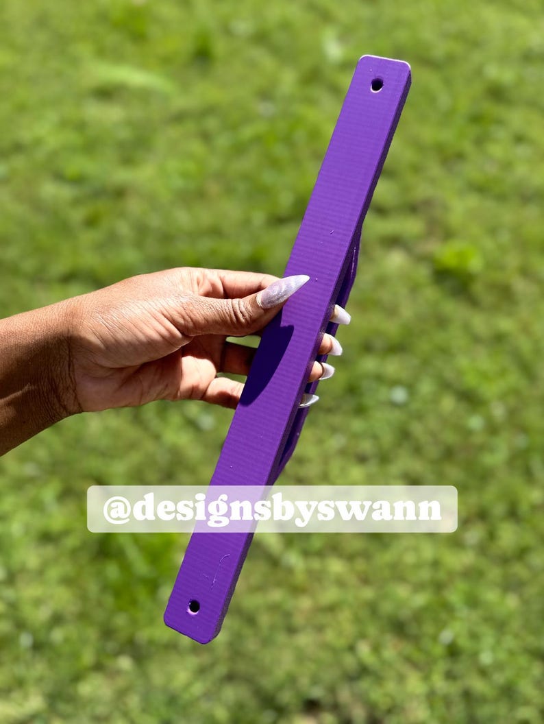May include: A long, rectangular purple object with a textured surface and two holes, held against a green background. The text "@designsbyswann" is visible on the object. The object appears to be a ruler or a similar measuring tool.
