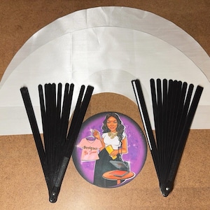 May include: Two black folding fans with white handles, and three white fabric circles, one on top of the other, with a round sticker in the center featuring a woman with a pink shirt and a purple background.