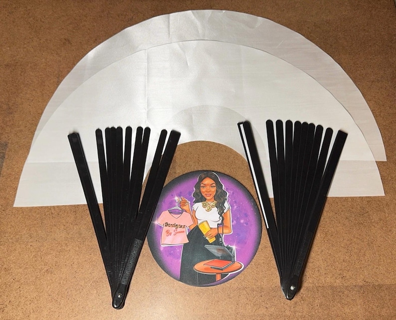 May include: Two black folding fans with white handles, and three white fabric circles, one on top of the other, with a round sticker in the center featuring a woman with a pink shirt and a purple background.