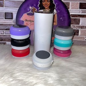 May include: A white tumbler with a black lid next to six colorful portable Bluetooth speakers. The speakers have a white base and a silicone outer ring in various colors, including purple, gray, turquoise, and pink.