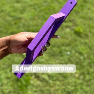 May include: A purple wooden object with a handle and a rectangular frame. The object has a hole at the end of the handle. The text "@designsbyswann" is on the object.