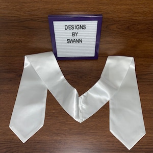 May include: White satin graduation stole with a pointed end. The stole is folded in half and laid flat on a wooden surface. A small sign with the text "DESIGNS BY SWANN" is visible in the background.