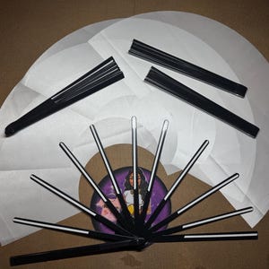 May include: A set of black folding fans with white paper panels. The fans are arranged in a circle with the handles pointing outwards. The fans are decorated with a white stripe down the center of each panel.