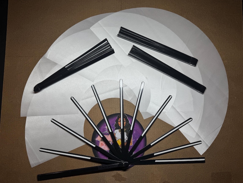 May include: A set of black folding fans with white paper panels. The fans are arranged in a circle with the handles pointing outwards. The fans are decorated with a white stripe down the center of each panel.