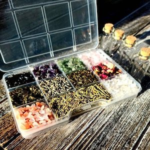 May include: A clear plastic storage box with 12 compartments filled with various herbs, crystals, and dried flowers. The box is open and the contents are visible. There are also several small glass bottles with cork stoppers.