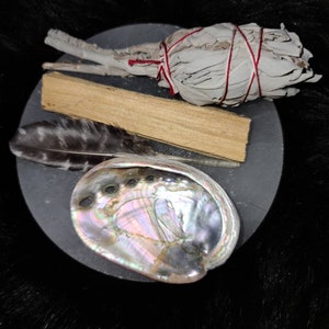 May include: A white sage smudge stick tied with red string, a palo santo stick, a black and white feather, and an abalone shell on a black surface.