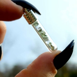 May include: A small glass vial filled with green and clear crystals, suspended from a silver chain. The vial is held between two fingers with black nail polish.