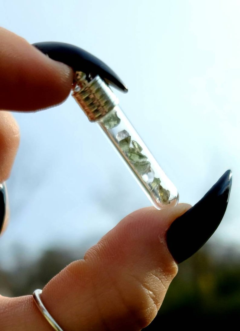 Op de afbeelding: Een klein glazen flesje gevuld met groene en heldere kristallen, opgehangen aan een zilveren ketting. Het flesje wordt tussen twee vingers met zwarte nagellak vastgehouden.
