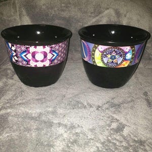 May include: Two black ceramic planters with colorful geometric designs. One planter features a purple and blue mandala design, while the other has a colorful design with a star and tree motif.