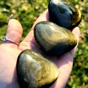 May include: Three golden sheen obsidian heart shaped stones. The stones are polished and have a smooth, shiny surface. The stones are a dark brown color with gold flecks.