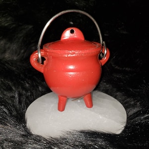May include: A small red cast iron cauldron with a handle and three legs. The cauldron is sitting on a white circular base.