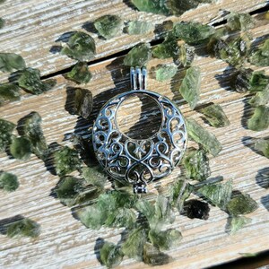 May include: A silver filigree cage pendant with a circular opening. The pendant is surrounded by small green gemstones on a wooden surface.