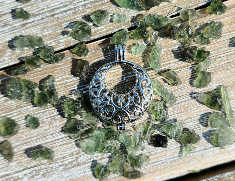 May include: A silver filigree cage pendant with a circular opening. The pendant is surrounded by small green gemstones on a wooden surface.