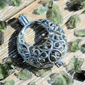 May include: A silver filigree charm with a circular opening, resembling a crescent moon. The charm is surrounded by small, green, translucent stones.