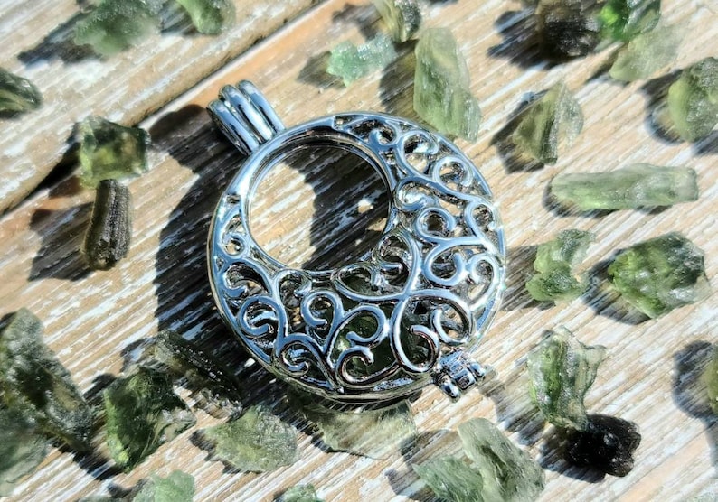 May include: A silver filigree charm with a circular opening, resembling a crescent moon. The charm is surrounded by small, green, translucent stones.