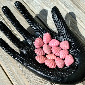 Puede incluir: Una colección de piedras pulidas en forma de corazón de color rosa en un plato negro con forma de mano.