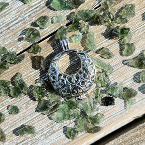 May include: A silver filigree cage pendant with a delicate, intricate design. The pendant is surrounded by small, green, translucent stones.