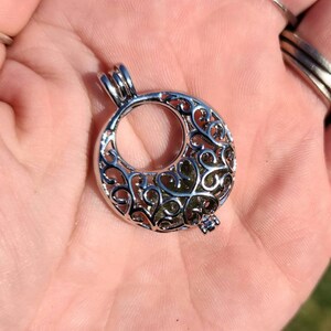 May include: Silver filigree charm with a circular opening. The charm has a delicate, intricate design with a heart-shaped pattern.