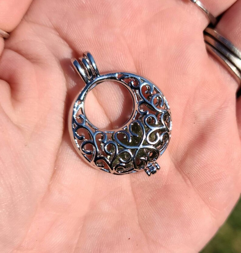 May include: Silver filigree charm with a circular opening. The charm has a delicate, intricate design with a heart-shaped pattern.