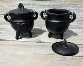 May include: Two black cast iron cauldrons with lids and three legs. The cauldron on the left has a lid with a handle and a decorative design on the side. The cauldron on the right has a lid with a handle and is smaller than the cauldron on the left.