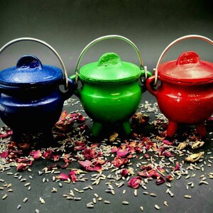 May include: Three small cast iron cauldrons with lids. The cauldrons are blue, green, and red. They are sitting on a surface covered in dried lavender and rose petals.