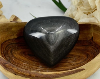 May include: A black heart-shaped obsidian crystal resting in a wooden bowl. The bowl is on a white marble surface.