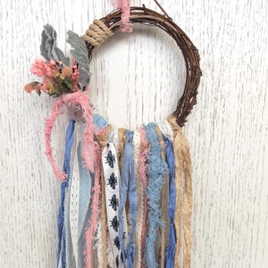 Peut inclure: Une couronne circulaire en vigne ornée d'un bouquet de fleurs séchées roses et de feuillage gris. Des bandes de tissu dans des tons bleu, rose, beige et blanc pendent de la couronne, incluant un ruban avec un motif d'abeilles noires imprimé.