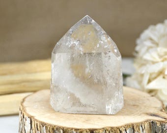May include: A clear, faceted quartz crystal point with a pointed top and a hexagonal base. The stone features internal inclusions and natural fractures that create a translucent, crystalline appearance with varying shades of white and gray.
